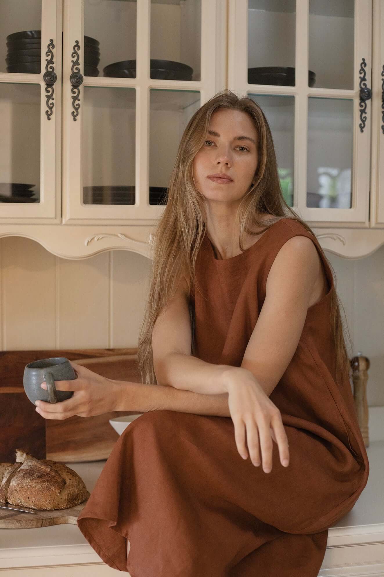 Woman wearing Cynthia Linen Dress Bark, sitting in a kitchen with a cup and bread, showcasing versatile style.