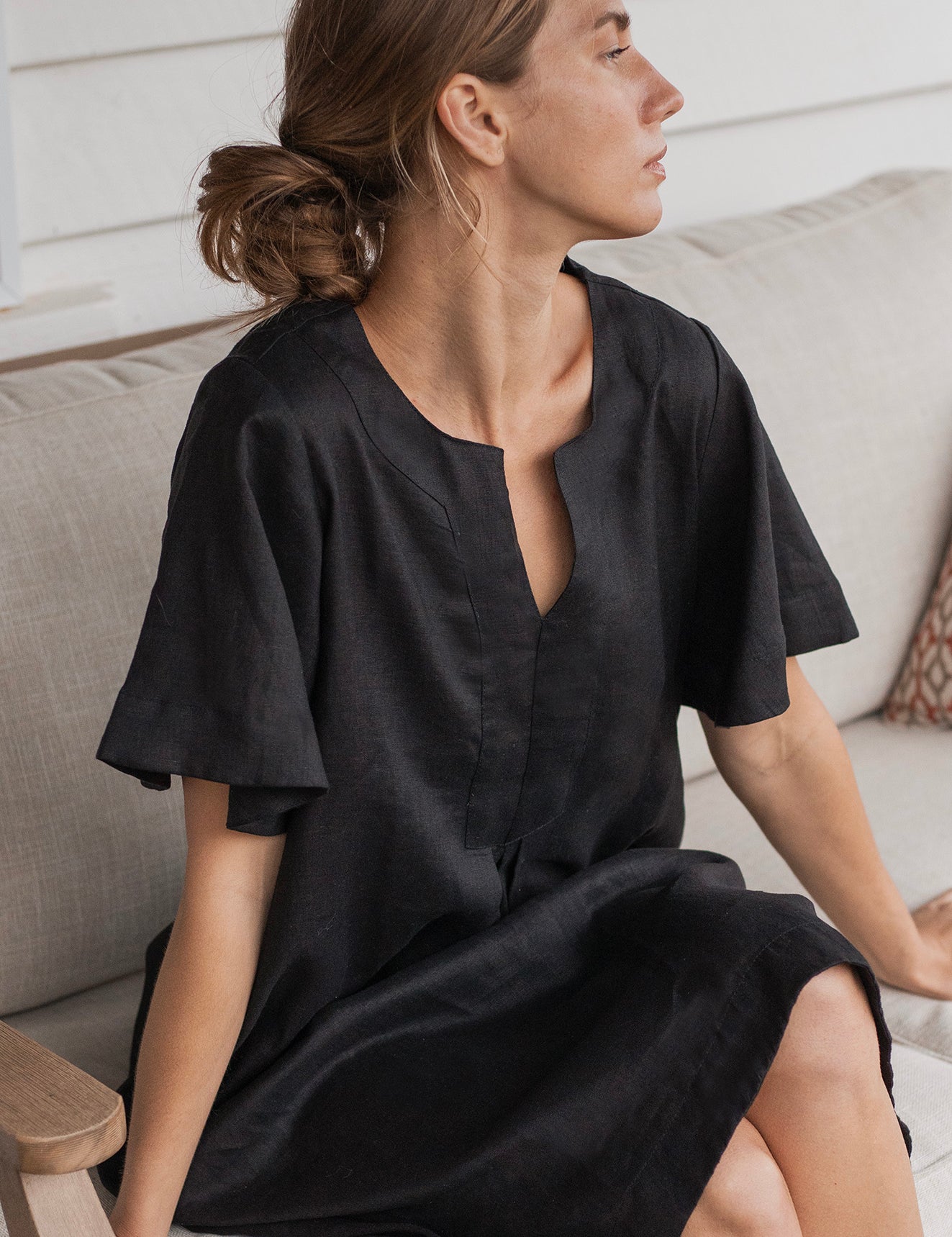 Charlotte Linen Dress Black worn by a model sitting on a couch, showcasing its effortless elegance and stylish design.