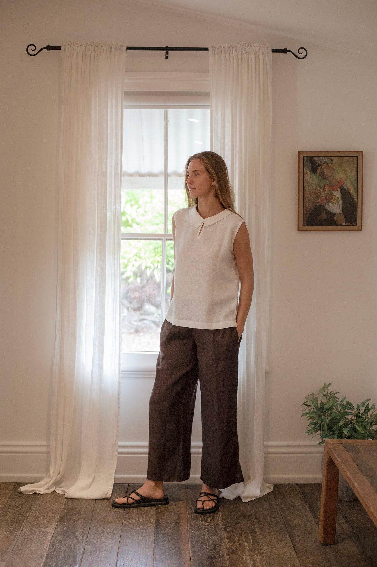 Woman wearing Paris Linen Pants Cocoa and a white top, standing by a window with sheer curtains.